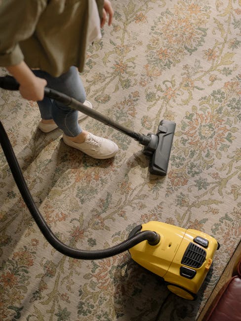 A person using a vacuum cleaner with a long hose attachment to clean a patterned area rug with floral motifs in a domestic setting. The rug features beige, green, and orange tones and covers a wooden floor. The individual is wearing beige sneakers, blue jeans, and a brown jacket, standing near the edge of the rug. The vacuum cleaner is a yellow, canister-style device placed on the floor, connected to the hose being directed across the carpet for deep cleaning. The room is well-lit, highlighting the cleanliness and detail of the rug and surrounding furniture, with hints of other home furnishings visible nearby. The image reflects professional carpet cleaning processes carried out by Carpet Cleaners Hammersmith, emphasizing surface cleaning and sanitisation in a domestic environment.