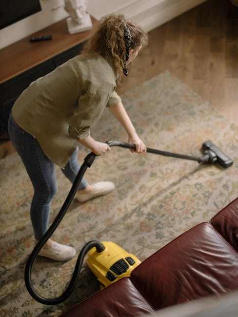 A woman with curly hair, wearing a beige jacket, dark jeans, and white shoes, is vacuuming a patterned area rug in a living room. She is using a yellow carpet cleaner with a black hose attachment, focusing on the carpet near a brown leather sofa. The room features hardwood flooring along the edges of the rug, a wooden sideboard with decorative items, and is well-lit with natural light. The scene illustrates a professional domestic cleaning process carried out by Carpet Cleaners Hammersmith, emphasizing surface cleaning and deep cleaning techniques for maintaining hygiene and cleanliness.