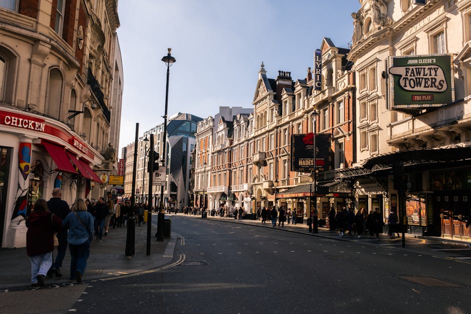 A busy street scene in the Lyric Theatre area of Hammersmith, London, featuring historic and modern buildings with ornate facades, signage for cinemas and shops, and pedestrians walking along the sidewalks. The street surface appears clean with visible road markings and streetlights illuminating the area. The storefronts include a mix of retail stores, theaters, and eateries, with some signs partially visible. Bright daylight highlights the architectural details and the bustling urban environment, reflecting the typical commercial and entertainment district where professional cleaning services like Carpet Cleaners Hammersmith ensure high standards of hygiene and surface cleanliness for both residential and commercial spaces in the area.