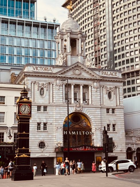The exterior of Victoria Palace Theatre in Hammersmith, featuring classical architectural details such as columns, statues, and decorative stonework on its facade. The building is surrounded by modern high-rise structures, with a prominent clock on a black and gold stand in front of the theatre. Pedestrians are visible walking along the pavement, some near the theatre entrance, and a white car is parked on the street. The scene is well-lit with daylight, and the overall appearance is clean and maintained. Carpet Cleaners Hammersmith offers expert domestic and commercial cleaning services, including surface and deep cleaning, to ensure hygienic environments in residential and theatre settings alike.