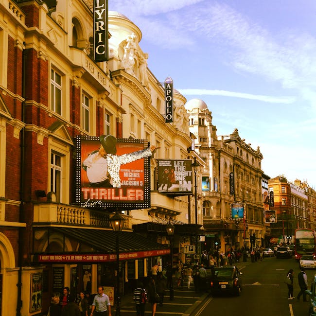 A busy street scene in the Lyric Theatre area of Hammersmith, London, featuring historic and modern buildings with ornate facades, signage for cinemas and shops, and pedestrians walking along the sidewalks. The street surface appears clean with visible road markings and streetlights illuminating the area. The storefronts include a mix of retail stores, theaters, and eateries, with some signs partially visible. Bright daylight highlights the architectural details and the bustling urban environment, reflecting the typical commercial and entertainment district where professional cleaning services like Carpet Cleaners Hammersmith ensure high standards of hygiene and surface cleanliness for both residential and commercial spaces in the area.