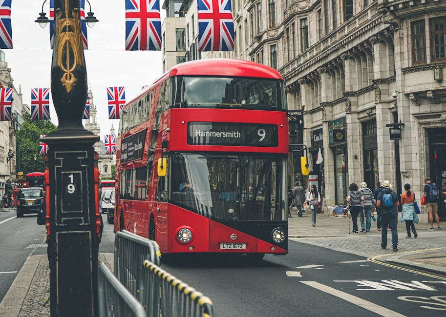 A red double-decker bus labeled 'Hammersmith 9' is traveling down a busy city street in London. The street is lined with historic, ornate buildings and is decorated with Union Jack flags hanging from 