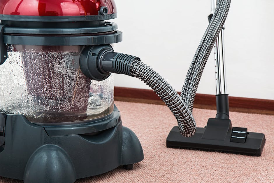 Close-up view of a wet vacuum cleaner with a flexible, patterned hose attached, positioned on a clean, pinkish-beige carpet in a residential interior. The vacuum's transparent dust container shows moisture and debris inside, indicating a deep cleaning process. The room features a wooden skirting board along the wall, and bright, even lighting highlights the cleanliness of the carpet surface. The image showcases equipment used by Carpet Cleaners Hammersmith for surface cleaning and hygiene maintenance, emphasizing professional domestic cleaning techniques in a home environment.