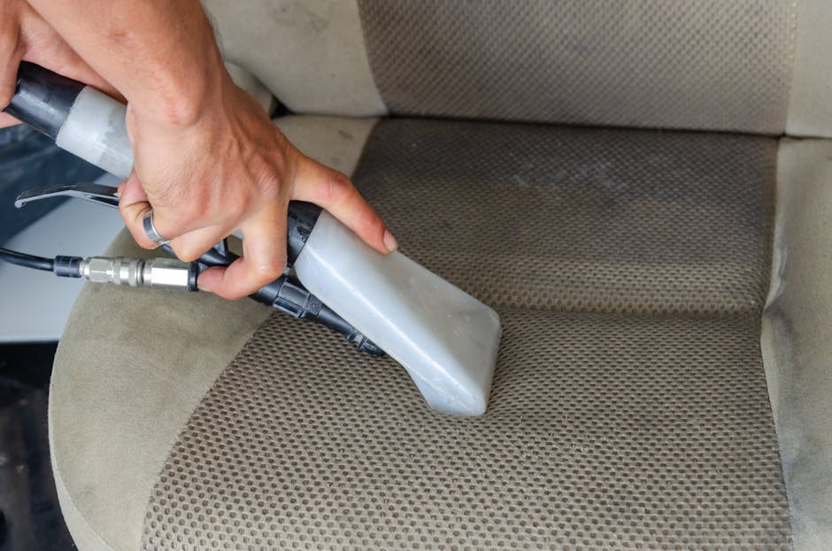 A close-up image showing a hand operating a handheld upholstery steam cleaner with a gray and black body, pressuring steam onto a beige fabric car seat with a textured, patterned surface. The seat appears to be in a vehicle interior, with visible dirt and stains being cleaned. The cleaning process is focused on deep surface cleaning and sanitisation of the upholstery, highlighting the use of professional tools by Carpet Cleaners Hammersmith for thorough hygiene maintenance of domestic or commercial seating areas.