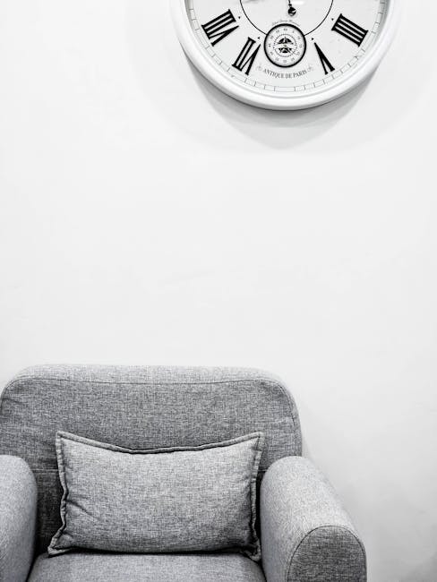 A close-up view of a grey upholstered armchair with a matching rectangular cushion placed on the seat. The chair is positioned against a plain white wall featuring a large, round, vintage-style clock with Roman numerals and black hands. The setting appears to be a clean, modern indoor living space with natural or soft lighting, emphasizing the tidy and well-maintained condition of the furniture. This scene illustrates the importance of surface cleaning and hygiene in domestic environments, which Carpet Cleaners Hammersmith offers as part of their upholstery cleaning services in the W6 Hammersmith area.